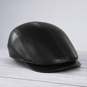 Puede incluir: Gorra plana de cuero negro con visera curva. La gorra tiene un acabado liso y brillante y está construida con múltiples paneles y costuras cosidas. La gorra está sobre una superficie de madera clara.