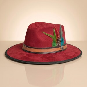 May include: A deep red fedora hat with a wide brim and a black edge. The hat features a tan leather band around the crown, accented with a silver-toned metal emblem and a cluster of green, red, and black-and-white spotted feathers on the side.