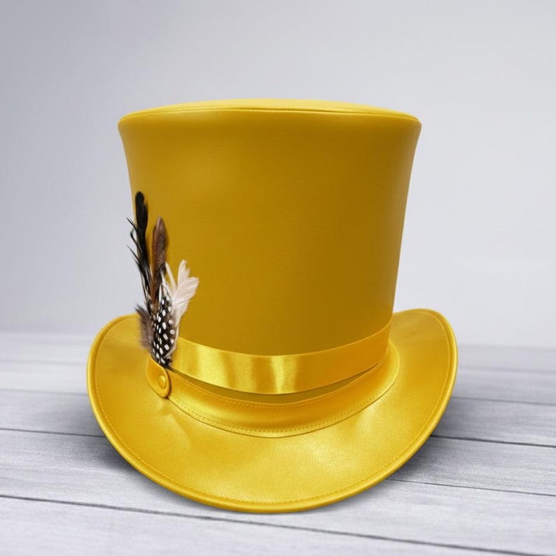 Puede incluir: Un sombrero de copa amarillo mostaza con ala ancha y cinta de raso. El sombrero est&aacute; adornado con un grupo de plumas en tonos marr&oacute;n, negro y blanco. Un dise&ntilde;o cl&aacute;sico.