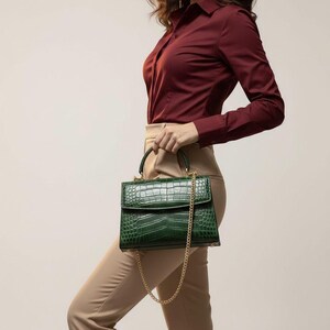May include: A green handbag with a crocodile skin effect and a gold chain strap. The bag has a top handle and a flap closure. The bag is held by a person wearing a burgundy button-down shirt and beige trousers.