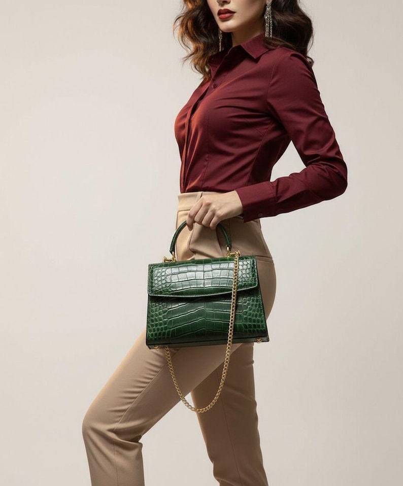 May include: A green handbag with a crocodile skin effect and a gold chain strap. The bag has a top handle and a flap closure. The bag is held by a person wearing a burgundy button-down shirt and beige trousers.