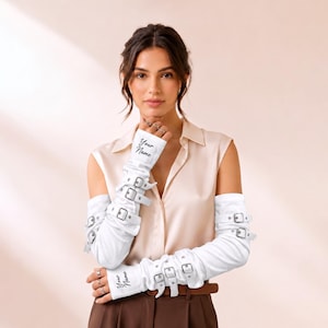 May include: A pair of white arm warmers featuring multiple silver-toned metal buckles and straps along the length of each sleeve. The fabric includes the black text "Your Name" printed on the upper and lower sections of the arm warmers.