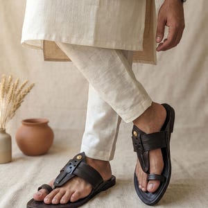 May include: A pair of dark brown leather sandals featuring a toe-ring design, textured straps, and small gold-toned circular accents. The sandals are worn with off-white linen trousers and a matching long-sleeved tunic top.
