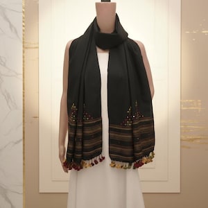 May include: A black scarf featuring horizontal stripes in shades of brown and gold, accented with small embroidered geometric patterns in red, green, and yellow. The bottom edges are finished with a row of small, multi-colored fabric tassels.