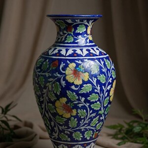 Könnte beinhalten: Eine blaue Keramikvase mit handgemaltem Blumenmuster aus gelben Blüten, grünen Blättern und weißen Ranken. Die Vase hat einen bauchigen Körper, einen schmalen Hals und einen ausgestellten Rand sowie dekorative geometrische Bordüren an Fuß und Hals.
