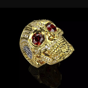 Puede incluir: Un anillo de calavera dorado con ojos de piedras preciosas rojas e incrustaciones de pequeños diamantes. El anillo tiene detalles intrincados y un acento plateado en el lateral. El anillo de calavera es una pieza audaz.