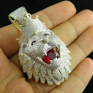 May include: A detailed pendant featuring a lion's head with a crown. The pendant is encrusted with small, sparkling stones, with a ruby-colored gem in the mouth. The lion's mane and crown have gold accents.