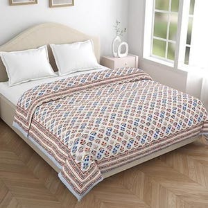Könnte beinhalten: Ein Bett mit einer weißen Matratze und zwei weißen Kissen. Eine gemusterte Decke mit einem sich wiederholenden Blumendesign in Rot, Blau und Beige bedeckt das Bett. Die Decke hat einen gestreiften Rand und einen hellblauen Rand.