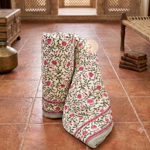 May include: A folded cotton quilt featuring a cream-coloured base with a repeating pattern of pink flowers and green vines. The fabric includes a border with thin horizontal stripes and a small circular logo with the text AMINOR printed on it.