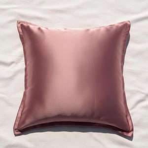 May include: A square, blush-coloured satin pillow with a subtle sheen. The pillow has a piped edge and rests on a white sheet. The pillow's colour is a muted rose, adding a touch of elegance to any bedroom decor.