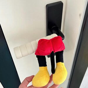 May include: A novelty door handle cover in the shape of Mickey Mouse's legs. The cover has red shorts, black legs, and yellow shoes. It's attached to a black door handle on a white door.