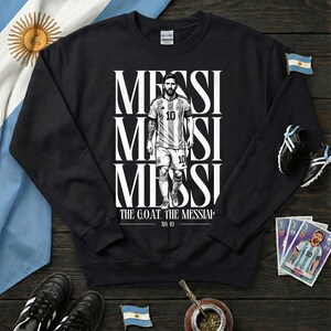 May include: Black sweatshirt with a graphic of a soccer player, with the name "MESSI" in white. The player is wearing a light blue and white jersey with the number 10. The text "THE G.O.A.T. THE MESSIAP" is below the image.