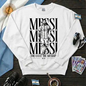 May include: White sweatshirt with a black and white illustration of a soccer player, with the name "MESSI" stacked vertically. The text "THE G.O.A.T. THE MESSIAH" and "NO. 10" are printed on the sweatshirt. The Argentine flag is visible.