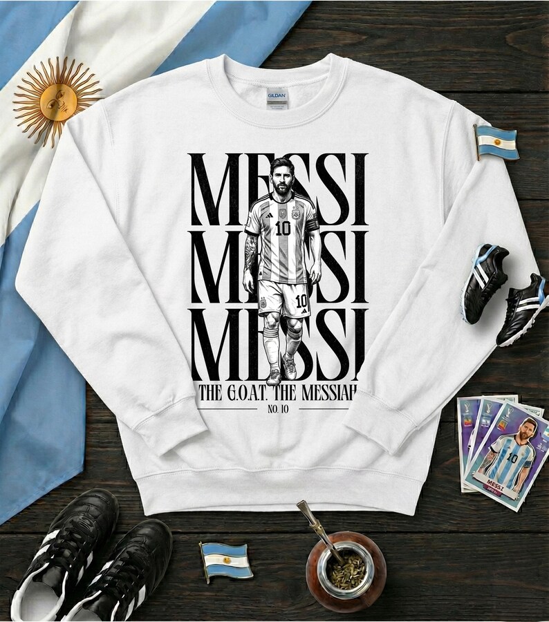 May include: White sweatshirt with a black and white illustration of a soccer player, with the name "MESSI" stacked vertically. The text "THE G.O.A.T. THE MESSIAH" and "NO. 10" are printed on the sweatshirt. The Argentine flag is visible.