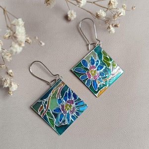 Pode incluir: Um par de brincos quadrados feitos à mão com um design floral. Os brincos apresentam um padrão floral azul, rosa e verde sobre um fundo turquesa. Os brincos são feitos com ganchos de prata.