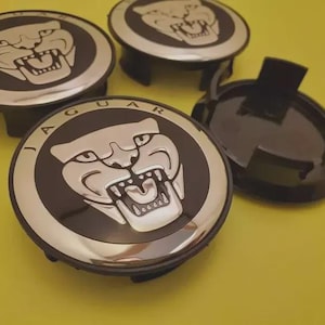 May include: Four black and silver Jaguar wheel center caps. Each cap features the Jaguar logo, a silver snarling jaguar head on a black background, surrounded by a silver ring with the word "JAGUAR". The caps are on a yellow background.