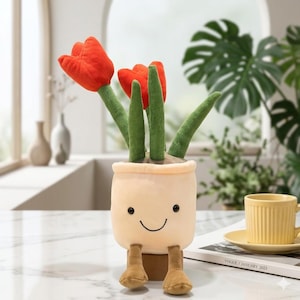 May include: A plush toy featuring a cream-colored pot with a smiling face and dangling brown feet, containing three bright orange fabric tulips with textured green stems and leaves. The soft, stuffed plant sits upright on a smooth surface.