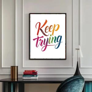 Op de afbeelding: Een ingelijste kunstafdruk met de woorden "Keep Trying" in een kleurrijk, cursief lettertype. Het kunstwerk staat tegen een witte achtergrond en is zwart omlijst. De print wordt weergegeven boven een zwart bureau met een teal stoel.
