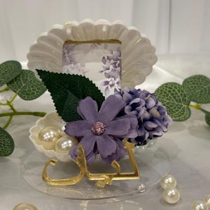 May include: A decorative centerpiece with a white, scalloped frame holding a floral print, decorated with purple flowers, green leaves, and faux pearls. A gold-coloured Arabic script is placed in front.