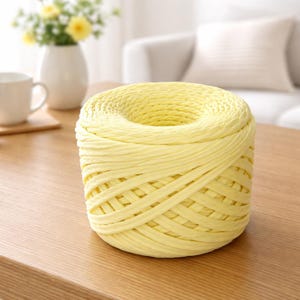 May include: A spool of pale yellow raffia yarn sits on a wooden table. The yarn is tightly wound, with a hole in the center. The background includes a white vase with yellow flowers and a white mug.