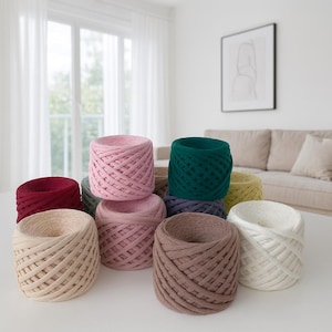 May include: A collection of colorful spools of thick, woven yarn. The yarn is in various shades, including pink, green, beige, and burgundy. The spools are arranged on a white surface, with a window and a couch in the background.