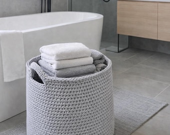 May include: A gray, crocheted storage basket filled with folded white and gray towels sits on a gray rug in a modern bathroom. A white bathtub and wooden vanity are in the background. The basket has two handles.