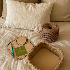 May include: A craft scene featuring a light brown woven basket, a wooden base with a circular design, a ball of brown yarn, and a teal crochet hook. A small green card is also present.