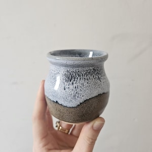 May include: A small, handcrafted ceramic pot with a rounded shape. The pot features a two-tone design, with a dark brown base and a light blue and white speckled upper section. The rim is a darker blue.