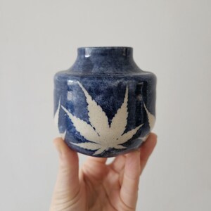 May include: A small, round, blue ceramic vase with a white leaf design. The vase is held in a hand against a white background. The vase has a dark blue glaze with white leaf patterns around the body.