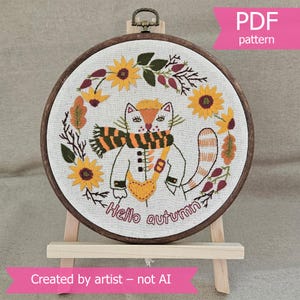 May include: Embroidered hoop art featuring a cat dressed in autumn attire, surrounded by sunflowers and fall foliage. The cat wears a scarf and a heart-shaped bag. Text reads "Hello autumn". The hoop is displayed on a small wooden easel.