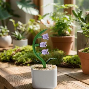 May include: A glass lily of the valley sculpture in a white square pot. The green stem curves gracefully, supporting three lavender-colored bell-shaped flowers. The pot sits on a wooden table, surrounded by other potted plants.