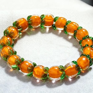 May include: A bracelet with orange and clear glass beads. The oval-shaped beads have green leaf accents. The beads are strung together in a circular design. The bracelet is displayed on a white surface.
