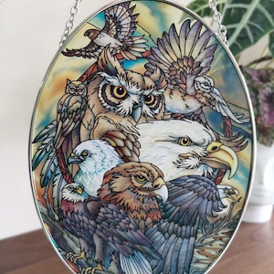 Puede incluir: Panel ovalado de vidrieras con una colección de aves rapaces, incluyendo búhos, águilas y halcones. La obra de arte muestra representaciones detalladas de las aves con colores vibrantes y patrones de plumas intrincados. Una cadena plateada está adjunta para colgar.
