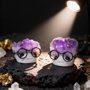 May include: Two whimsical amethyst crystal sculptures, each adorned with small black glasses and a smiling face. The crystals are set on a dark, textured surface, with a spotlight illuminating them. Additional crystals and gold-colored pyrite are scattered around.