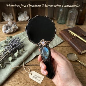 Op de afbeelding: Een handspiegel met een cirkelvormig zwart obsidiaan glazen oppervlak en een handvat versierd met een grote, iriserende blauwe labradoriet edelsteen en een klein hartvormig accent. De tekst Handcrafted Obsidian Mirror with Labradorite is zichtbaar.
