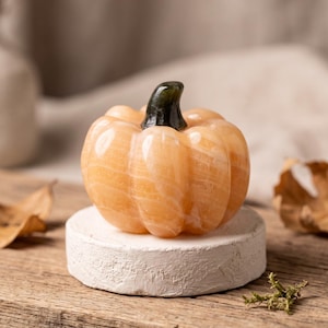 May include: A carved orange stone pumpkin figurine with a smooth, polished surface and a dark green stone stem. The pumpkin features vertical ridges and a rounded shape, resting on a circular, textured white base.