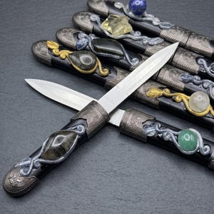 May include: A collection of letter openers with black handles, silver-toned metal accents, and various embedded gemstones including lapis lazuli, citrine, and green aventurine. Each handle features intricate metal scrollwork and a pointed blade.
