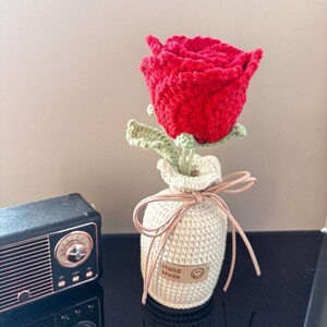 Könnte beinhalten: Eine handgefertigte rote Häkelrose mit grünen Blättern, präsentiert in einer beigefarbenen Häkelvase, die mit einem braunen Band gebunden ist. Die Vase hat ein kleines Etikett mit der Aufschrift "Hand Made" mit einem Smiley.