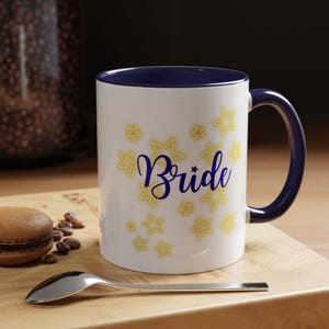Könnte beinhalten: Ein weißer Keramikbecher mit dunkelblauem Innenraum und Henkel. Die Außenseite zeigt das Wort Bride in dunkelblauer Schreibschrift, umgeben von mehreren gelb umrandeten Sternen in verschiedenen Größen, die auf der Fläche verteilt sind.