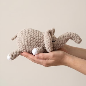 May include: A beige crocheted elephant toy made from soft, chunky yarn. The elephant features a rounded body, large floppy ears, a trunk, and small white accents on the tips of its feet. The texture is consistent and plush throughout the design.