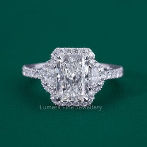 May include: A silver-toned ring featuring a large rectangular radiant-cut center stone surrounded by a halo of small round diamonds. Two half-moon shaped side stones are set on either side of the center, with a pavé diamond band. Lumera Fine Jewellery.