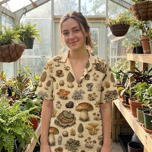 May include: A short-sleeve button-down shirt in a light cream color featuring an all-over print of various mushrooms and fungi in shades of brown, tan, and orange. The shirt has a classic collar, a straight hem, and a front button closure.