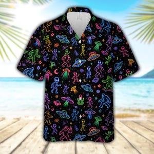 May include: A black short-sleeve button-down shirt featuring a repeating neon-style print of aliens, flying saucers, and Bigfoot figures. The design includes vibrant outlines in various colors against a dark background with a classic collar.