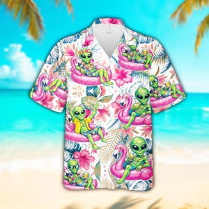 May include: Short-sleeved button-down shirt featuring a tropical pattern with green aliens relaxing on pink flamingo floats. The shirt has a white background with pink flowers and tropical leaves. The aliens are wearing sunglasses and holding drinks.