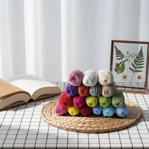 May include: A collection of multi-coloured yarn balls, including pink, purple, red, yellow, green, blue, white, and grey, stacked on a woven mat. A framed botanical print and an open book are also in view.