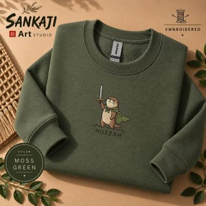 Pode incluir: Uma camisola de gola redonda verde musgo com um bordado de uma lontra castanha a usar uma capa verde e a segurar uma espada prateada. Abaixo da lontra, a palavra HUZZAH está bordada com fio escuro. A peça tem gola e punhos canelados.