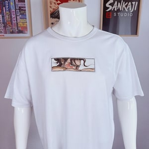May include: White t-shirt with a rectangular embroidered design featuring a manga-style illustration of a person with brown hair and red markings. The shirt is displayed on a white mannequin. The background includes framed Japanese art and a sign that says "Sankaji Studio."