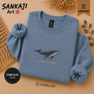 May include: A stone blue crewneck sweatshirt featuring an embroidered design of a whale swimming beneath a crescent moon and stars. The text "WE RIDE AT DAWN" is embroidered in white below the whale. The garment has long sleeves with ribbed cuffs.