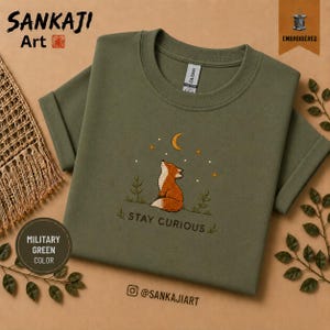 Op de afbeelding: Een militair groen t-shirt met een geborduurd ontwerp van een vos die naar een maansikkel en sterren kijkt. Onder de vos is de tekst STAY CURIOUS geborduurd in een donkere draad. Het t-shirt heeft een ronde hals en korte mouwen.