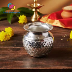 May include: A silver-colored, decorative pot with a diamond pattern on the lower half and a textured rim. The pot sits on a dark wooden surface, with yellow flowers and a brass lamp in the background. The text "G.G.S. Enterprise" is visible.